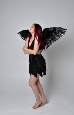 Siyah elbise ve tüylü melek kanatları takan kızıl saçlı bir kızın tam boy portresi. Stüdyo geçmişine karşı poz vermek.