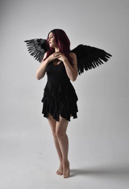 Siyah elbise ve tüylü melek kanatları takan kızıl saçlı bir kızın tam boy portresi. Stüdyo geçmişine karşı poz vermek.