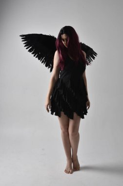 Siyah elbise ve tüylü melek kanatları takan kızıl saçlı bir kızın tam boy portresi. Stüdyo geçmişine karşı poz vermek.