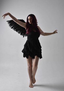 Siyah elbise ve tüylü melek kanatları takan kızıl saçlı bir kızın tam boy portresi. Stüdyo geçmişine karşı poz vermek.