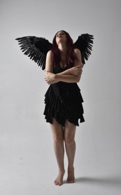 Siyah elbise ve tüylü melek kanatları takan kızıl saçlı bir kızın tam boy portresi. Stüdyo geçmişine karşı poz vermek.
