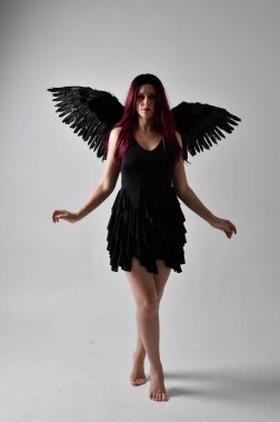 Siyah elbise ve tüylü melek kanatları takan kızıl saçlı bir kızın tam boy portresi. Stüdyo geçmişine karşı poz vermek.