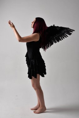 Siyah elbise ve tüylü melek kanatları takan kızıl saçlı bir kızın tam boy portresi. Stüdyo geçmişine karşı poz vermek.
