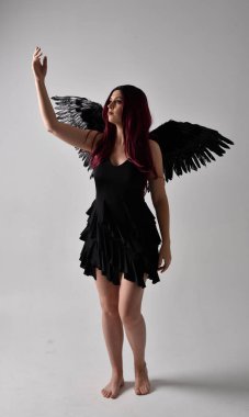 Siyah elbise ve tüylü melek kanatları takan kızıl saçlı bir kızın tam boy portresi. Stüdyo geçmişine karşı poz vermek.