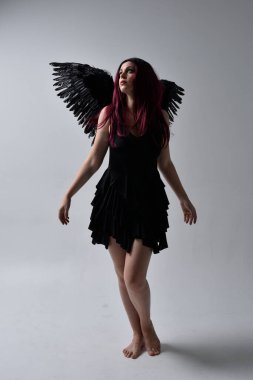 Siyah elbise ve tüylü melek kanatları takan kızıl saçlı bir kızın tam boy portresi. Stüdyo geçmişine karşı poz vermek.