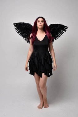Siyah elbise ve tüylü melek kanatları takan kızıl saçlı bir kızın tam boy portresi. Stüdyo geçmişine karşı poz vermek.