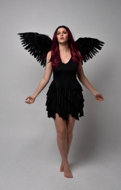 Siyah elbise ve tüylü melek kanatları takan kızıl saçlı bir kızın tam boy portresi. Stüdyo geçmişine karşı poz vermek.