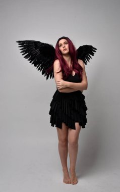 Siyah elbise ve tüylü melek kanatları takan kızıl saçlı bir kızın tam boy portresi. Stüdyo geçmişine karşı poz vermek.