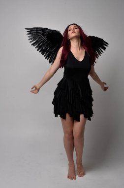 Siyah elbise ve tüylü melek kanatları takan kızıl saçlı bir kızın tam boy portresi. Stüdyo geçmişine karşı poz vermek.