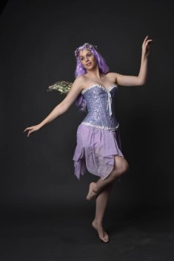 Peri kanatları ve çiçek tacıyla fantezi korse giyen mor saçlı bir kızın tam boy portresi. Karanlık bir stüdyo geçmişine karşı poz vermek.