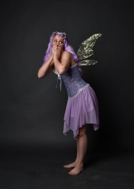 Peri kanatları ve çiçek tacıyla fantezi korse giyen mor saçlı bir kızın tam boy portresi. Karanlık bir stüdyo geçmişine karşı poz vermek.