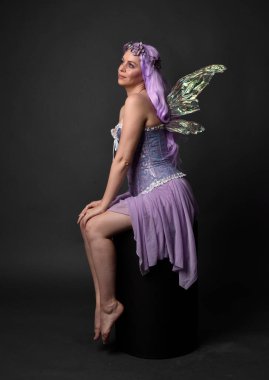 Peri kanatları ve çiçek tacıyla fantezi korse giyen mor saçlı bir kızın tam boy portresi. Stüdyonun arka planında otururken.