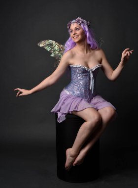 Peri kanatları ve çiçek tacıyla fantezi korse giyen mor saçlı bir kızın tam boy portresi. Stüdyonun arka planında otururken.