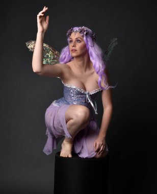 Peri kanatları ve çiçek tacıyla fantezi korse giyen mor saçlı bir kızın tam boy portresi. Stüdyonun arka planında otururken.