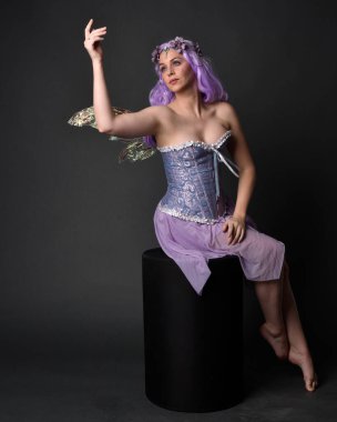 Peri kanatları ve çiçek tacıyla fantezi korse giyen mor saçlı bir kızın tam boy portresi. Stüdyonun arka planında otururken.