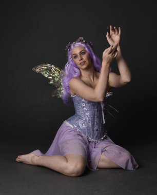 Peri kanatları ve çiçek tacıyla fantezi korse giyen mor saçlı bir kızın tam boy portresi. Stüdyonun arka planında otururken.