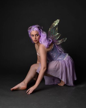Peri kanatları ve çiçek tacıyla fantezi korse giyen mor saçlı bir kızın tam boy portresi. Stüdyonun arka planında otururken.