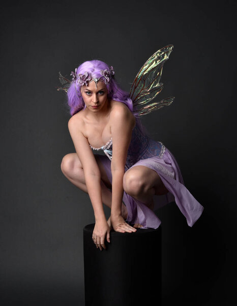 Full length portrait of a purple haired girl wearing fantasy corset dress with fairy wings and flower crown.  Seated pose against a dark studio background.