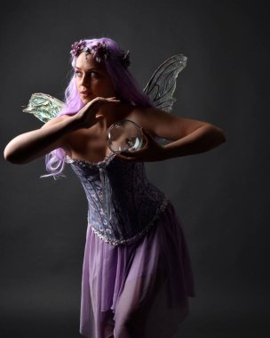 Peri kanatları ve çiçek tacıyla süslü fantezi korse giyen mor saçlı bir kızın portresi. Gölgeli ışıklandırmalı karanlık bir stüdyonun arka planında poz vermek..