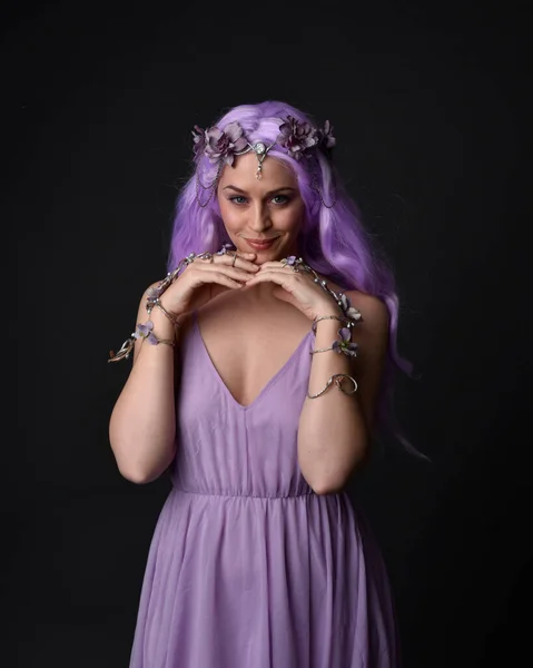 Close up portrait of a purple haired  girl wearing fantasy corset dress with fairy wings and flower crown, casting a spell.  Posing against a dark studio background.