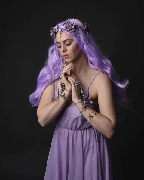 Close up portrait of a purple haired  girl wearing fantasy corset dress with fairy wings and flower crown, casting a spell.  Posing against a dark studio background.