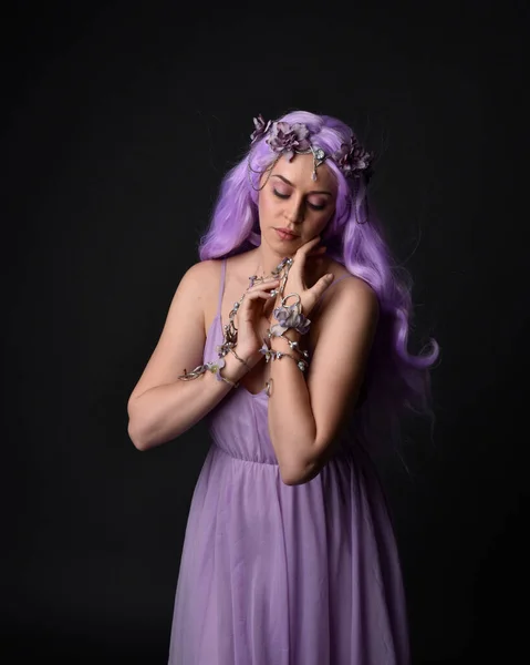 Close up portrait of a purple haired  girl wearing fantasy corset dress with fairy wings and flower crown, casting a spell.  Posing against a dark studio background.