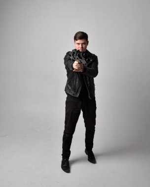 Deri ceket giyen ve elinde bilim kurgu silahı tutan esmer bir adamın tam boy portresi. Gri bir stüdyonun arka planında ayakta durarak poz vermek.