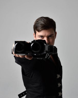 Deri ceket giyen ve elinde bilim kurgu silahı tutan esmer bir adamın portresi. Eylem, gri bir stüdyonun arka planında izole edilmiş.