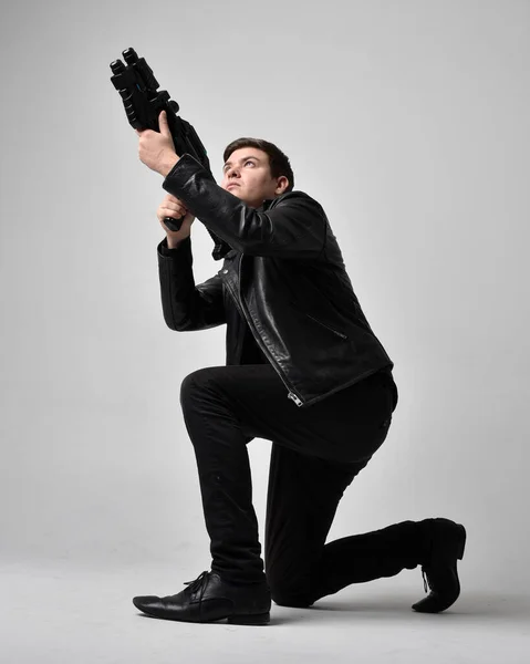 stock image Full length portrait of a  brunette man wearing leather jacket  and holding a science fiction gun.  kneeling  action pose isolated  against a grey studio background.