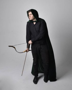 Siyah gömlek, yelek ve elinde ok ve yay tutan yeşil kadife pelerin giyen esmer bir adamın tam boy portresi. Gri bir stüdyonun arka planında ayakta durarak poz vermek.