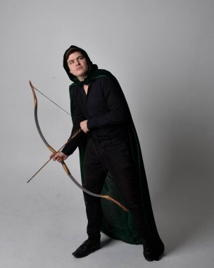 Full length portrait of a  brunette man wearing black shirt, waistcoat and a green velvet cloak holding a bow and arrow.  Standing  action pose isolated  against a grey studio background. clipart
