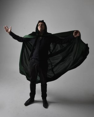 Siyah gömlek, yelek ve yeşil kadife pelerin giyen esmer bir adamın tam boy portresi. Gri bir stüdyonun arka planında ayakta durarak poz vermek.