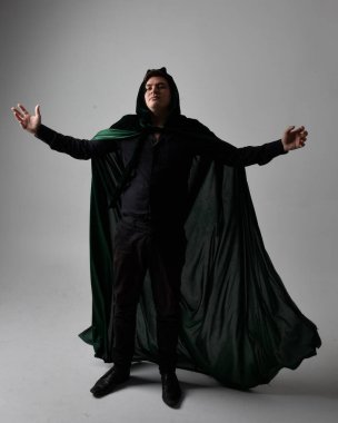 Siyah gömlek, yelek ve yeşil kadife pelerin giyen esmer bir adamın tam boy portresi. Gri bir stüdyonun arka planında ayakta durarak poz vermek.