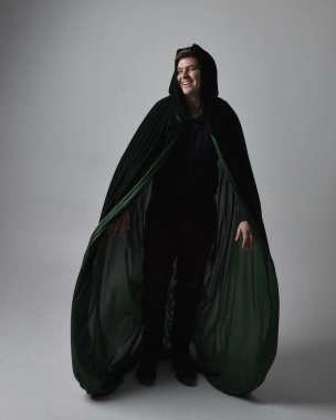 Siyah gömlek, yelek ve yeşil kadife pelerin giyen esmer bir adamın tam boy portresi. Gri bir stüdyonun arka planında ayakta durarak poz vermek.