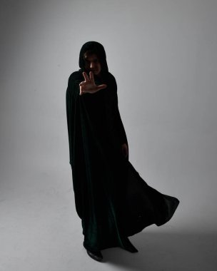 Siyah gömlek, yelek ve yeşil kadife pelerin giyen esmer bir adamın tam boy portresi. Gri bir stüdyonun arka planında ayakta durarak poz vermek.