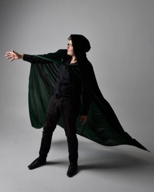Full length portrait of a  brunette man wearing black shirt, waistcoat and a green velvet cloak.  Standing  action pose isolated  against a grey studio background. clipart