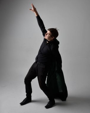 Siyah gömlek, yelek ve yeşil kadife pelerin giyen esmer bir adamın tam boy portresi. Gri bir stüdyonun arka planında ayakta durarak poz vermek.