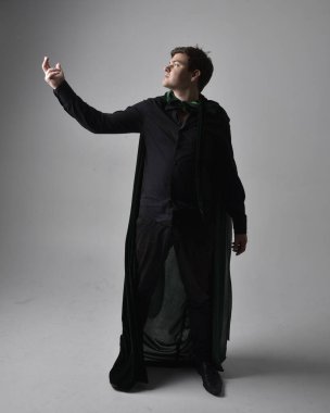 Siyah gömlek, yelek ve yeşil kadife pelerin giyen esmer bir adamın tam boy portresi. Gri bir stüdyonun arka planında ayakta durarak poz vermek.