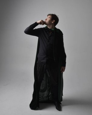 Siyah gömlek, yelek ve yeşil kadife pelerin giyen esmer bir adamın tam boy portresi. Gri bir stüdyonun arka planında ayakta durarak poz vermek.