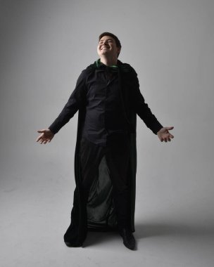 Siyah gömlek, yelek ve yeşil kadife pelerin giyen esmer bir adamın tam boy portresi. Gri bir stüdyonun arka planında ayakta durarak poz vermek.