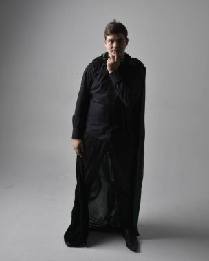 Siyah gömlek, yelek ve yeşil kadife pelerin giyen esmer bir adamın tam boy portresi. Gri bir stüdyonun arka planında ayakta durarak poz vermek.