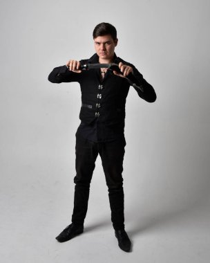 Siyah gömlek giyen esmer bir adamın uzun bir portresi ve elinde kılıç tutan gotik bir yelek. Gri bir stüdyonun arka planında ayakta durarak poz vermek.