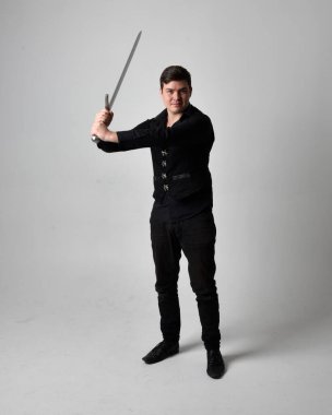 Siyah gömlek giyen esmer bir adamın uzun bir portresi ve elinde kılıç tutan gotik bir yelek. Gri bir stüdyonun arka planında ayakta durarak poz vermek.