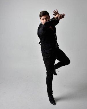 Siyah gömlek giyen esmer bir adamın uzun bir portresi ve elinde kılıç tutan gotik bir yelek. Gri bir stüdyonun arka planında ayakta durarak poz vermek.