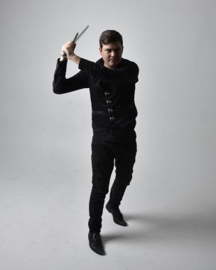 Siyah gömlek giyen esmer bir adamın uzun bir portresi ve elinde kılıç tutan gotik bir yelek. Gri bir stüdyonun arka planında ayakta durarak poz vermek.