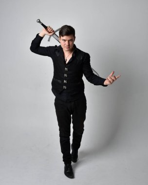 Siyah gömlek giyen esmer bir adamın uzun bir portresi ve elinde kılıç tutan gotik bir yelek. Gri bir stüdyonun arka planında ayakta durarak poz vermek.