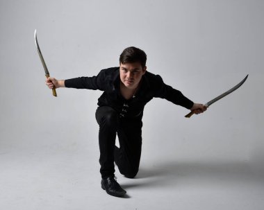 Siyah gömlek giyen esmer bir adamın uzun bir portresi ve elinde kılıç tutan gotik bir yelek. Gri bir stüdyonun arka planında ayakta durarak poz vermek.