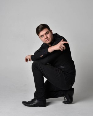 Siyah gömlek ve gotik yelek giyen esmer bir adamın tam boy portresi. Gri bir stüdyonun arka planında izole bir poz..