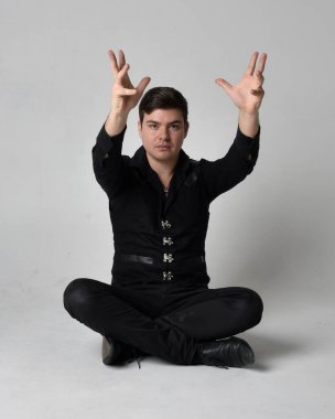 Siyah gömlek ve gotik yelek giyen esmer bir adamın tam boy portresi. Gri bir stüdyonun arka planında izole bir poz..