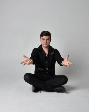 Siyah gömlek ve gotik yelek giyen esmer bir adamın tam boy portresi. Gri bir stüdyonun arka planında izole bir poz..